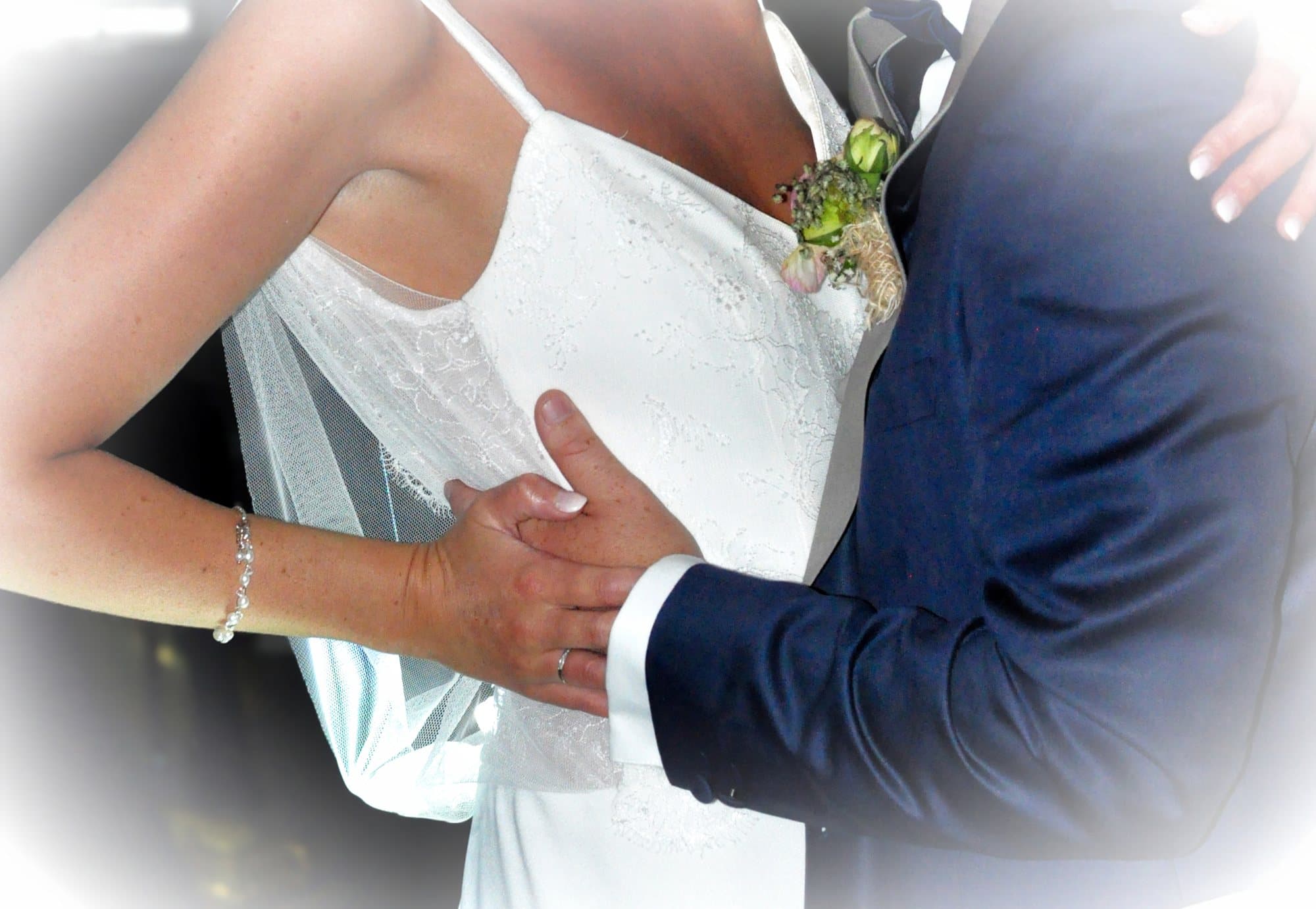 Gros plan sur la main du marié posée dans le dos de la mariée, boutonnière et alliances