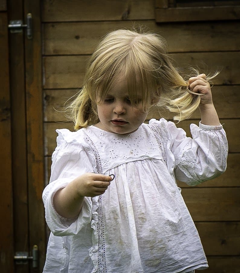 Petite fille blonde en robe blanche entortillant ses cheveux devant une porte en bois